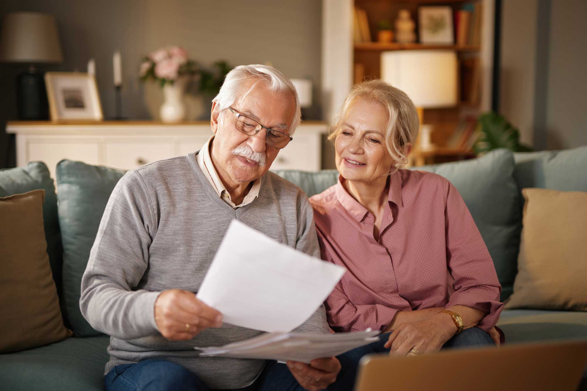An elderly man wearing glasses reads paperwork while sitting with his wife on a sofa